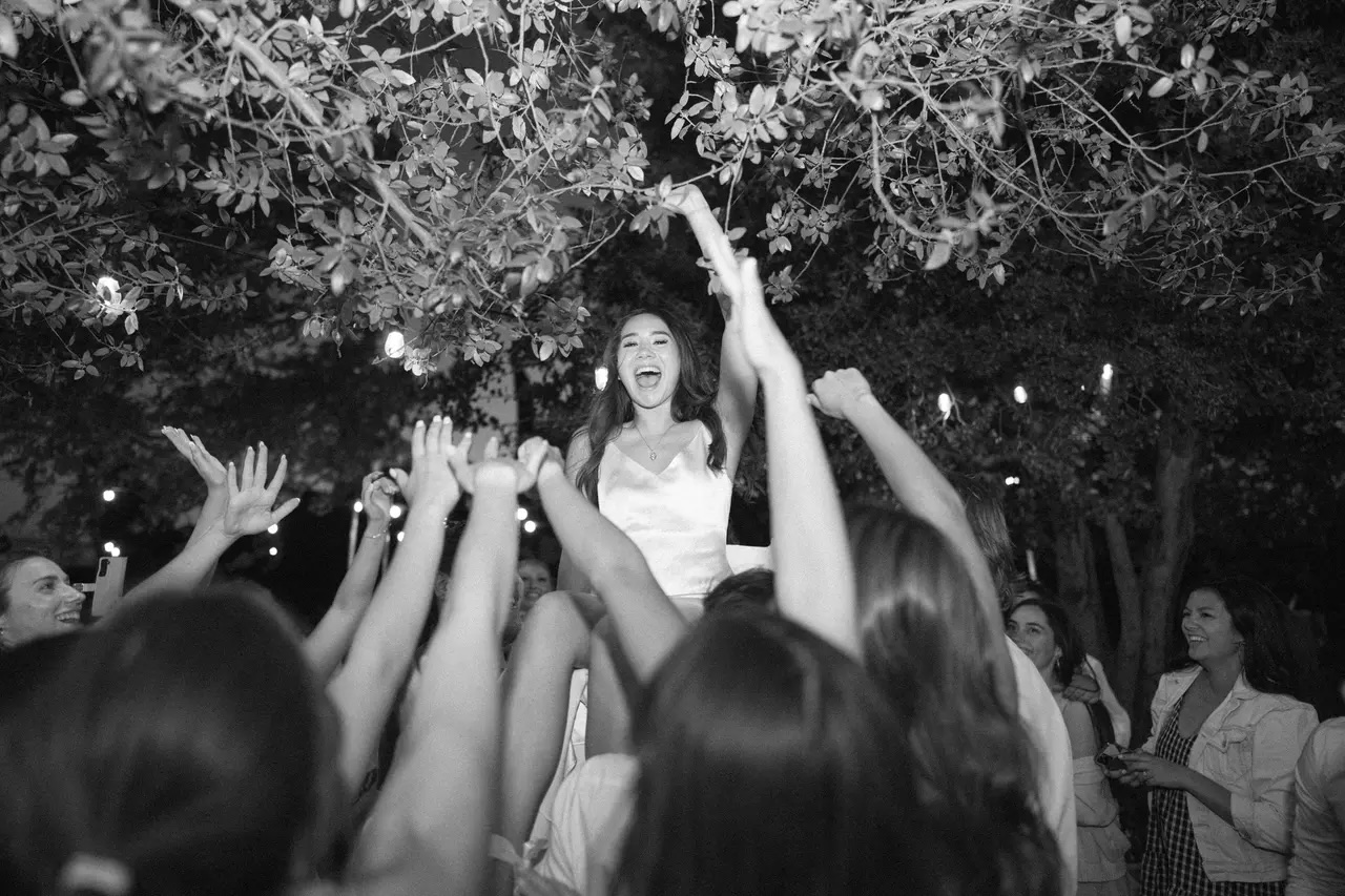 Black-and-white photo of a smiling bride lifted above the crowd on the dance floor, guests cheering with their hands raised under string lights